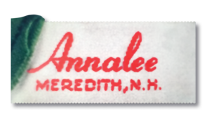 Satin tag with pinked edges from 1954-1955. White tag with red lettering script on first line with block lettering on second line. 